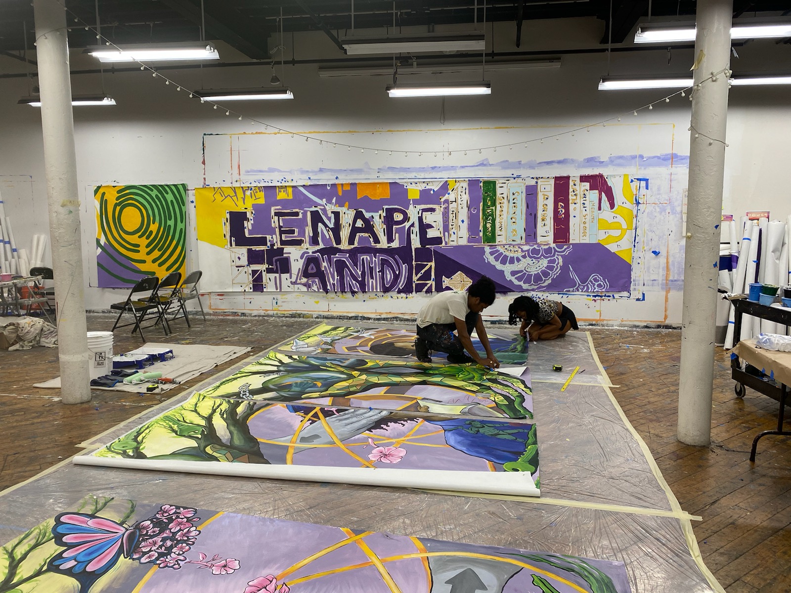 Two artists working on murals on the ground in Brooklyn Justice Initiatives in front of colorful illustration that reads "Lenape Land."