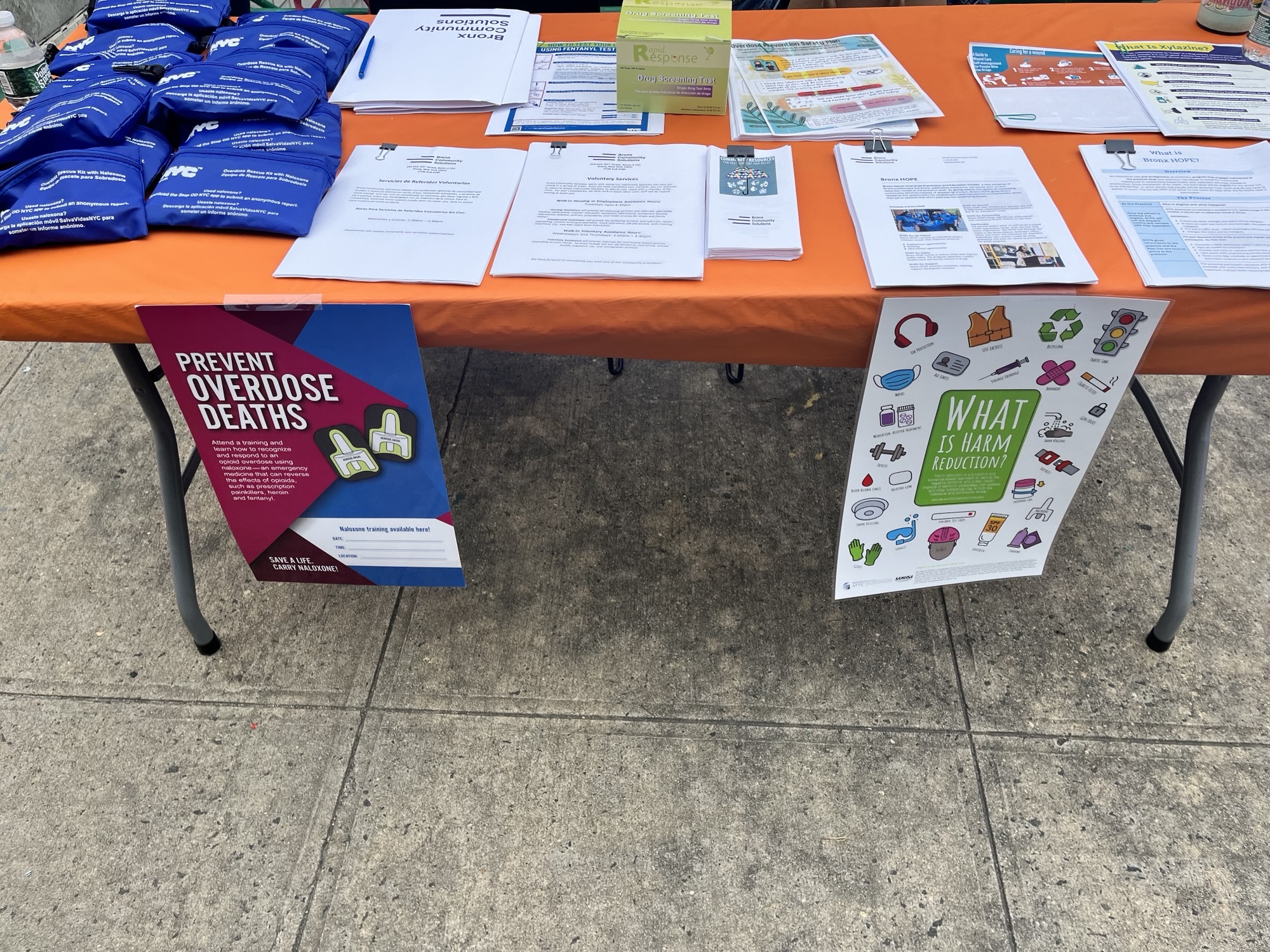 Outdoor table with overdose prevention kits, fentanyl testing strips, informational packets, and flyers reading "Prevent Overdose Deaths" and "What Is Harm Reduction?"