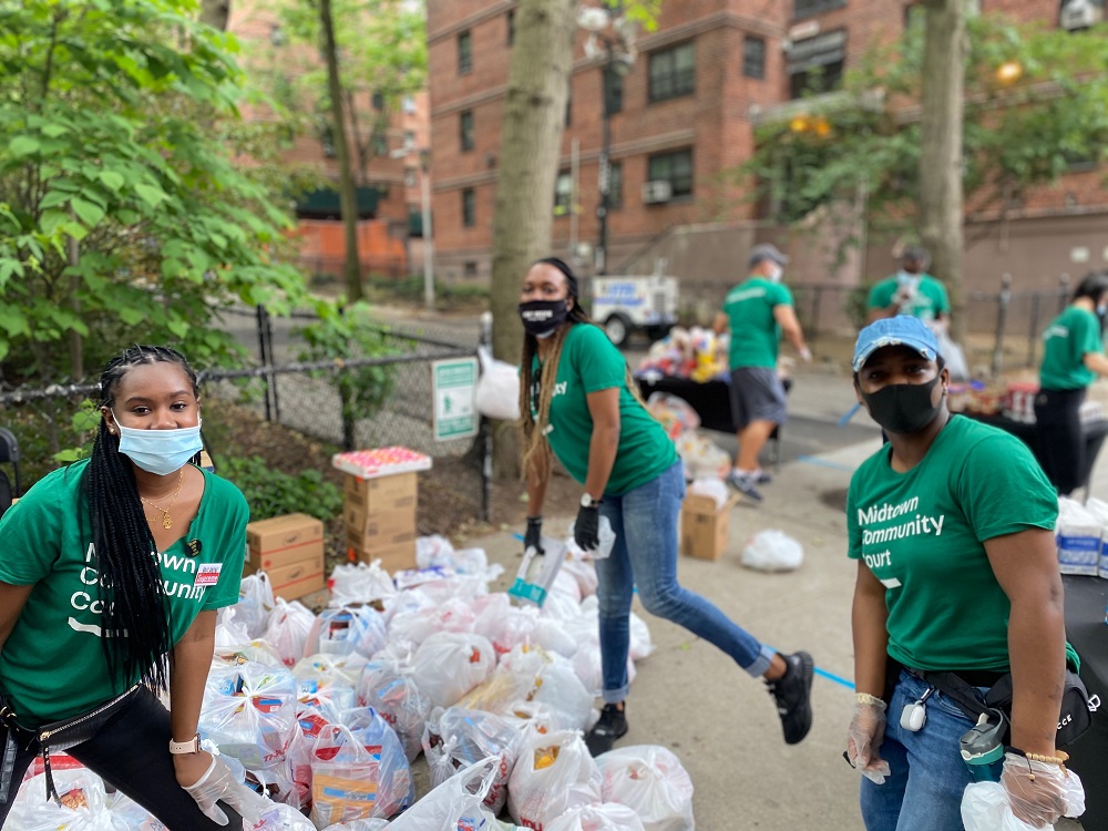 Three women in masks and green t-shirts that say Midtown Community Court distribute supplies to the community