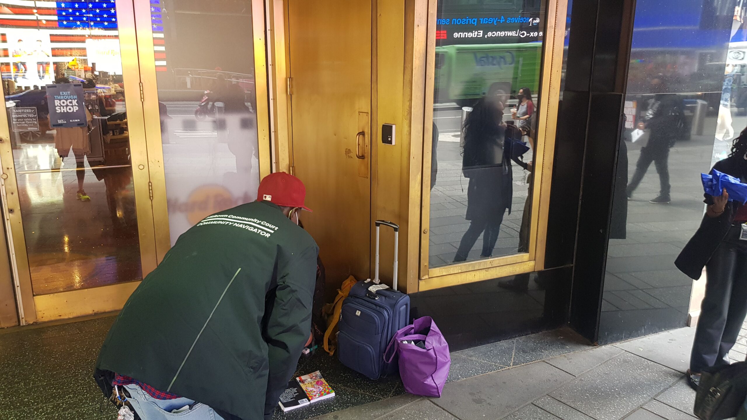 A community navigator in a green jacket and red hat bends over to talk to a person sitting on the sidewalk with their belongings