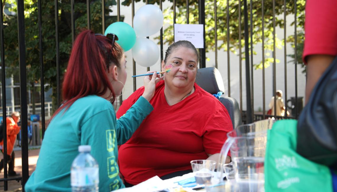 Midtown Community Court creates safe, thriving communities with events, like this community fair. Pic: woman in green shirt painting a multi-colored heart on the face of a woman in a red shirt, who is facing the camera