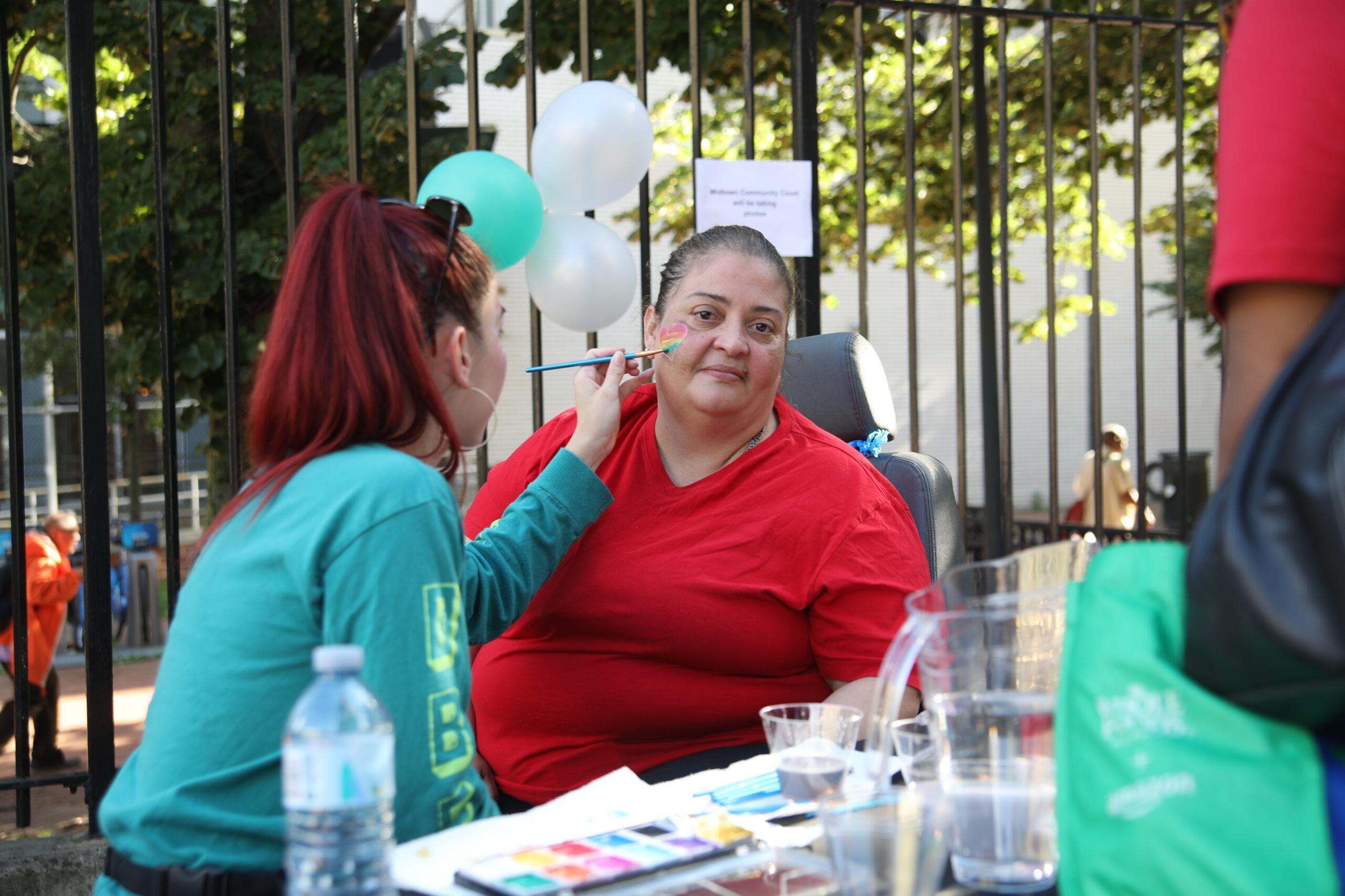 Midtown Community Court creates safe, thriving communities with events, like this community fair. Pic: woman in green shirt painting a multi-colored heart on the face of a woman in a red shirt, who is facing the camera