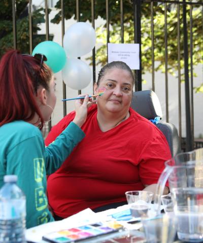 Midtown Community Court creates safe, thriving communities with events, like this community fair. Pic: woman in green shirt painting a multi-colored heart on the face of a woman in a red shirt, who is facing the camera