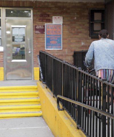 A Black, older resident of Wagner Houses uses her walker to get to the door of the public housing's building