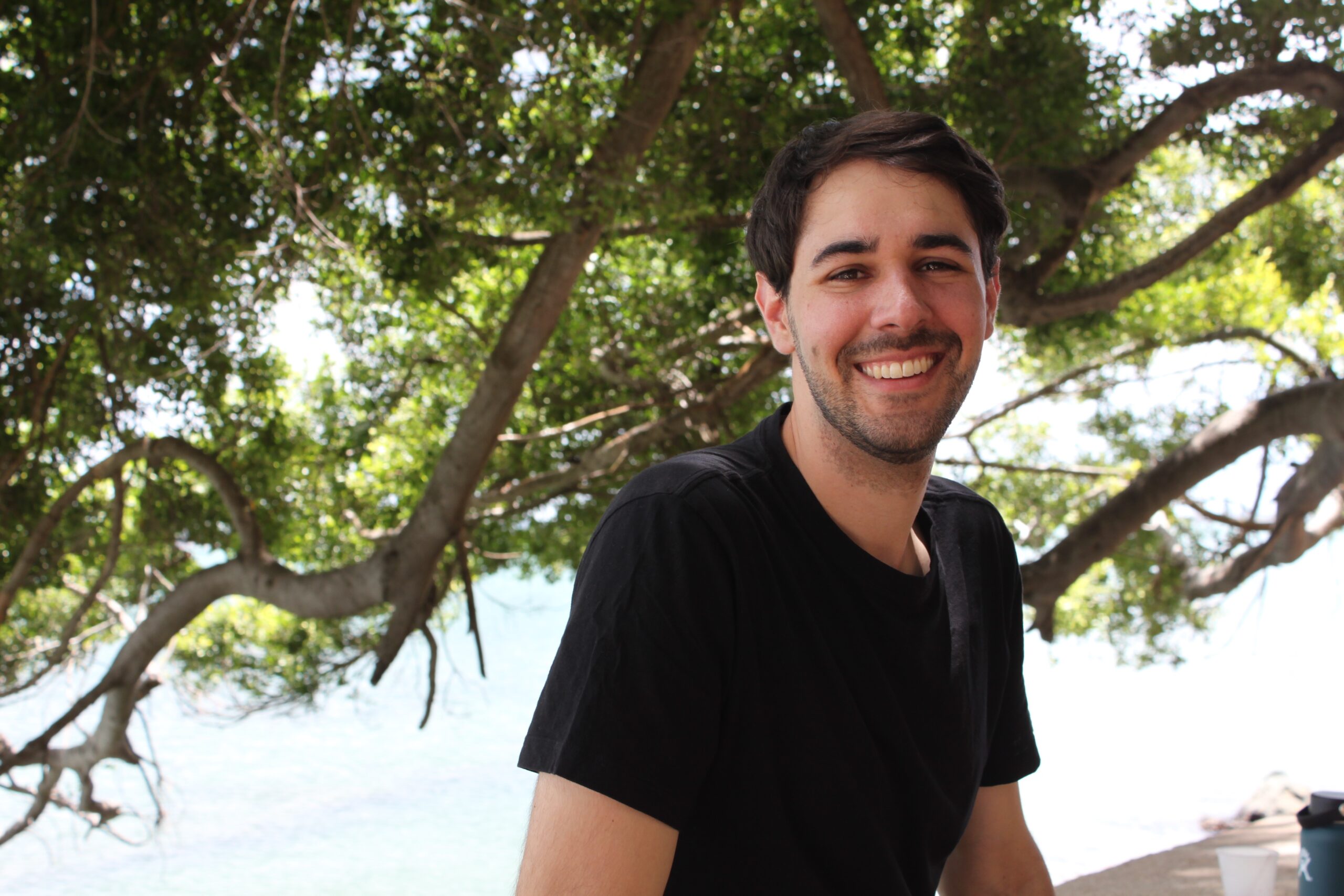 Joe Barrett outside by trees in a black tee shirt looking at the camera smiling