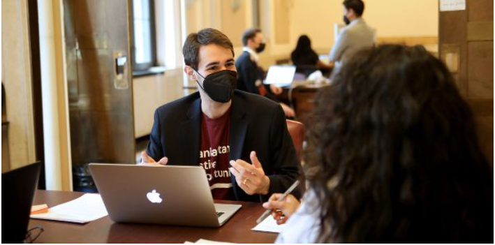 Manhattan Justice Opportunities staff member is facing the camera sitting at a table in front of a laptop in a maroon shirt speaking with a participant who has their back turned to the camera