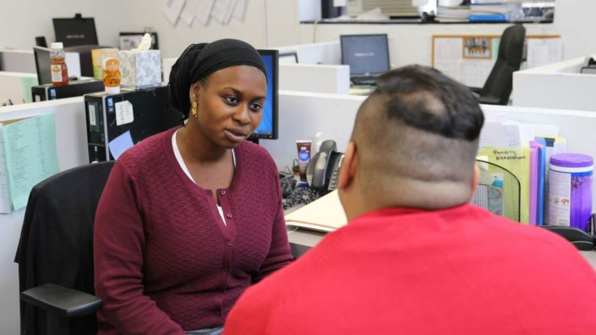 A staff member in a burgundy shirt is sitting and facing a participant talking in an office