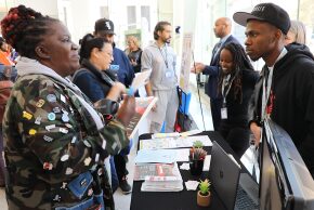 A young Black man speaks to a Black woman at the policy breakfast from a table with informational flyers.