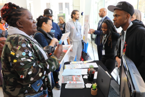 A young Black man speaks to a Black woman at the policy breakfast from a table with informational flyers.
