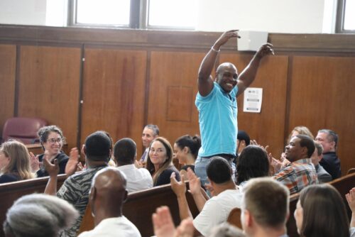 Participant celebrating with arms up in the courtroom aisle at an Alternatives to Incarceration graduation.
