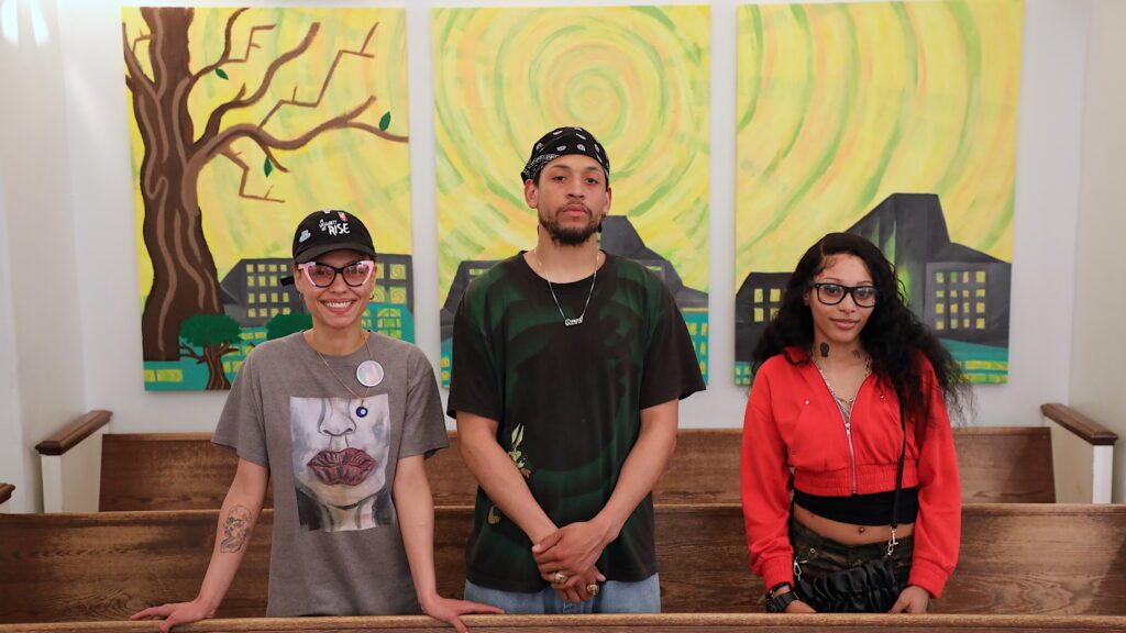 Three artists stand in front of three-panel painting in Midtown courtroom featuring trees, buildings, and a yellow spiral sky.