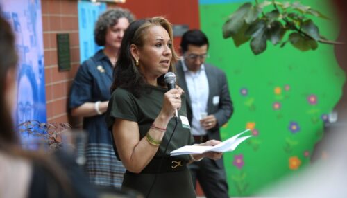 a woman in black (Rasmia Kirmani) speaking with a microphone in front of two people