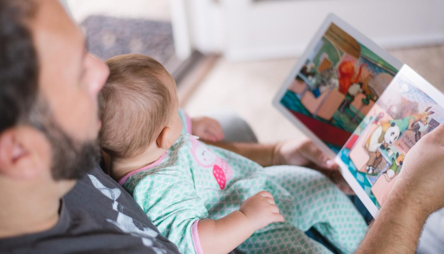 A caregiver with a small child in their lap holds a bright, colorful book open to read.