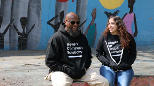 Man in Newark Community Solutions hoodie and woman wearing black jacket over Center for Justice Innovation T-shirt sit on a ledge in front of outdoor mural