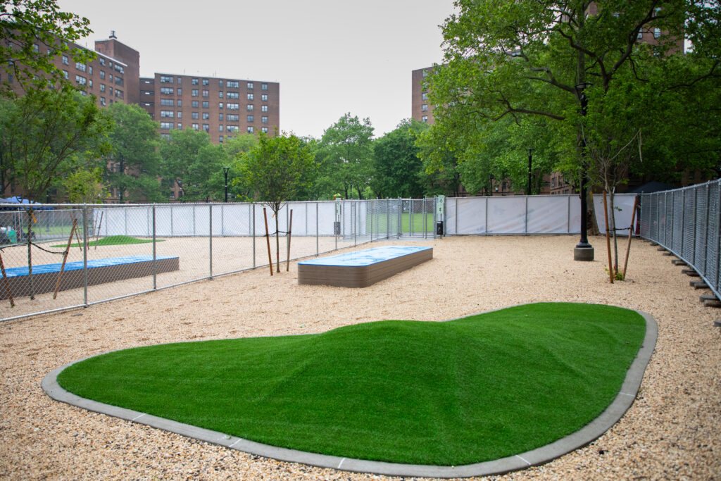 Gravel lot, elevated platform, turf mound, and trees inside gated area at Castle Hill dog park.