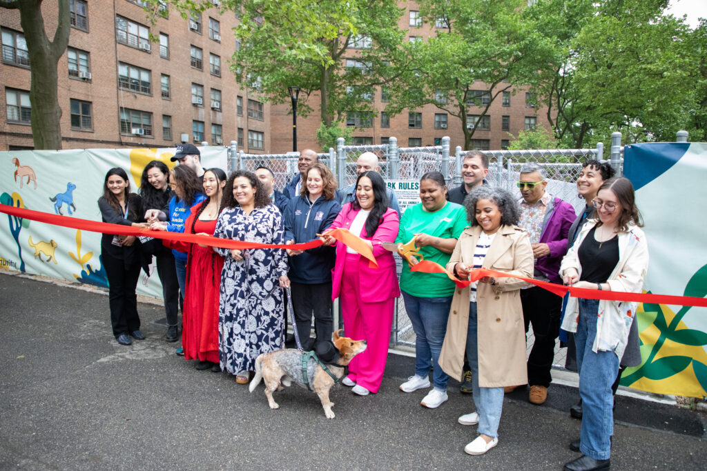 Dog wearing top hat looks up at group of people cutting a red ribbon to unveil the Castle Hill Houses' new dog park.