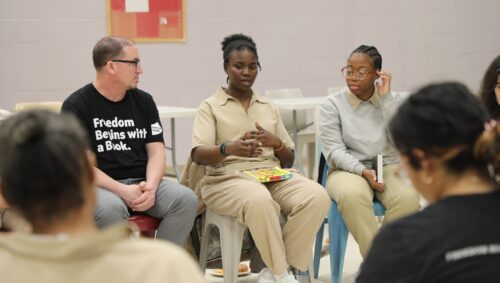 A woman sits in beige attire with a book on her lap discussing in a circle with other women and a man with a T-shirt that reads 