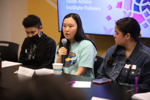 Three young people sit in a row. The center person holds a microphone and is mid-sentence.