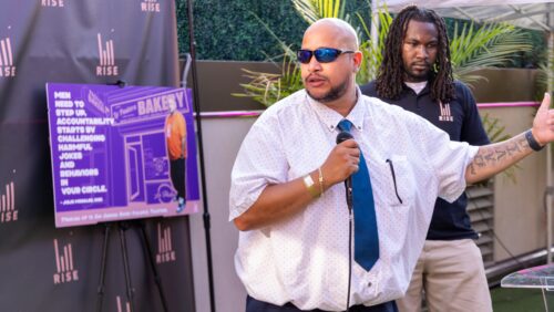 Man in shirt and tie with sunglasses stands with microphone in front of another man wearing a 