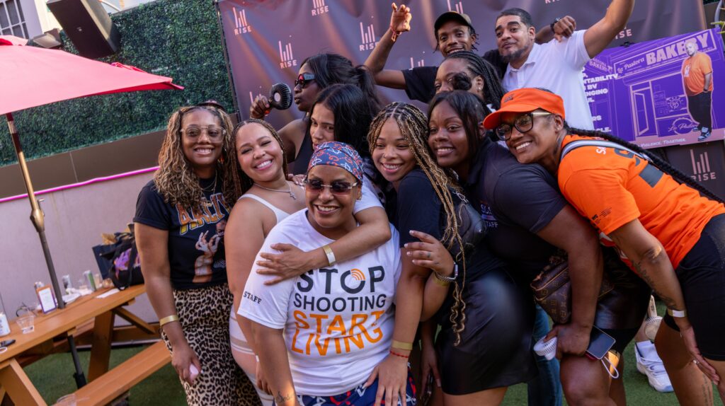 A group of women and men pose for a group picture at the Men Rising launch event; one woman in front wears a white T-shirt that reads "Stop shooting, start living."
