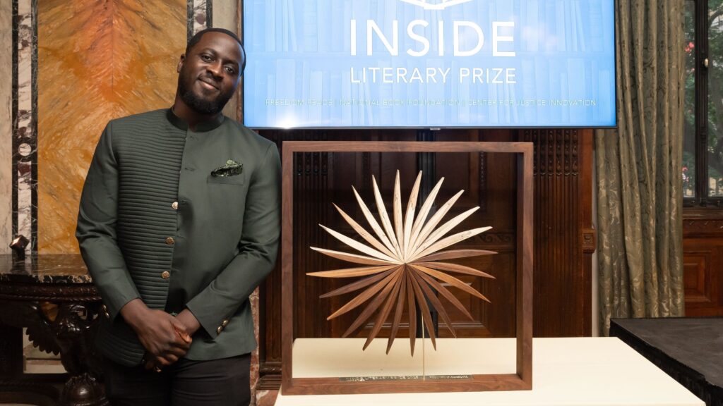 Author Nana Kwame Adjei-Brenyah stands next to the Inside Literary Prize award, a wooden display in the shape of book leaves fanning out into a circular formation.