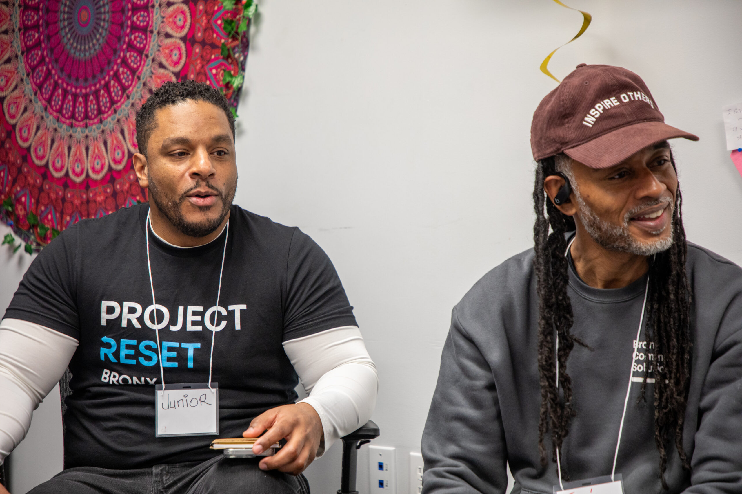 Two men in Project Reset and Bronx Community Solutions shirts sit in a circle in front of a circular colorful tapestry on the wall.