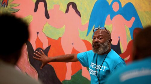 Man in blue Project Reset T-shirt speaks while pointing to a mural featuring three pink silhouetted figures in abstract style.