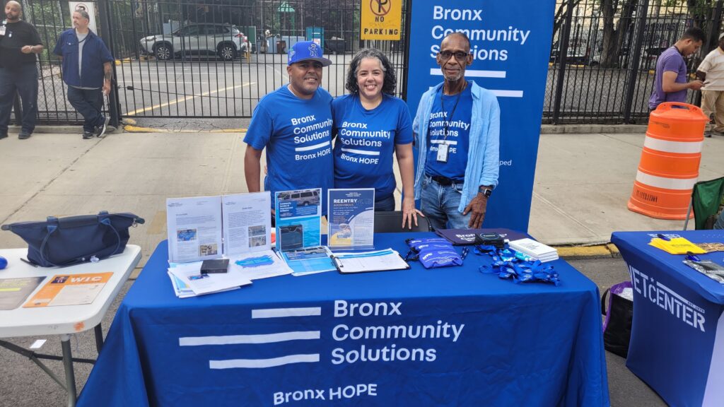 Three people in Bronx Community Solutions shirts stand at a table outside with flyers, pamphlets, and pouches.
