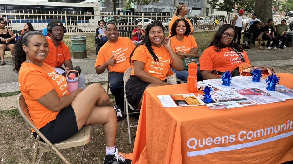 Group of people with orange "Queens Community Justice Center" T-shirts sit smiling at a table with pamphlets and resources