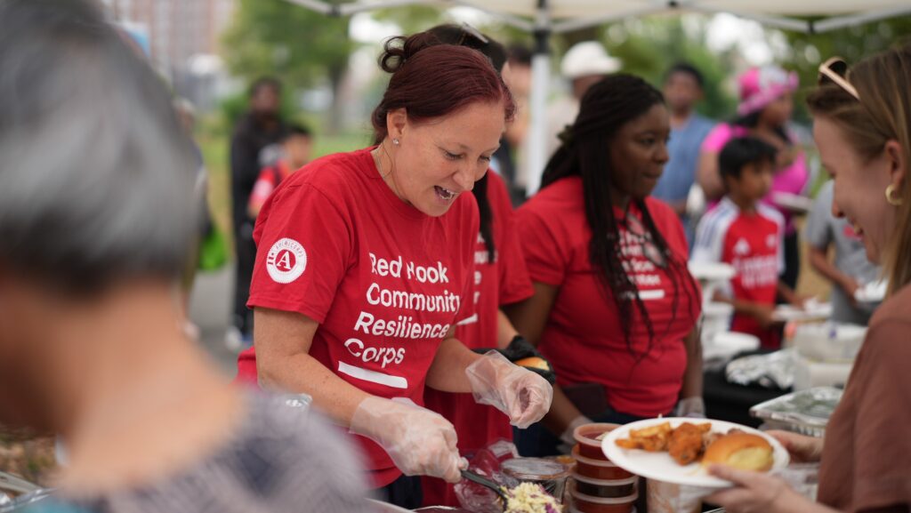 Woman wearing red "Red Hook Community Resilience Corps" T-shirt serves food to attendees at National Night Out.