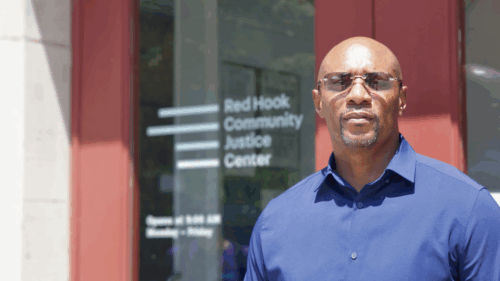 Close-up shot of Thaddeus Canty with blue button-down shirt and sunglasses standing at entrance to Red Hook Community Justice Center