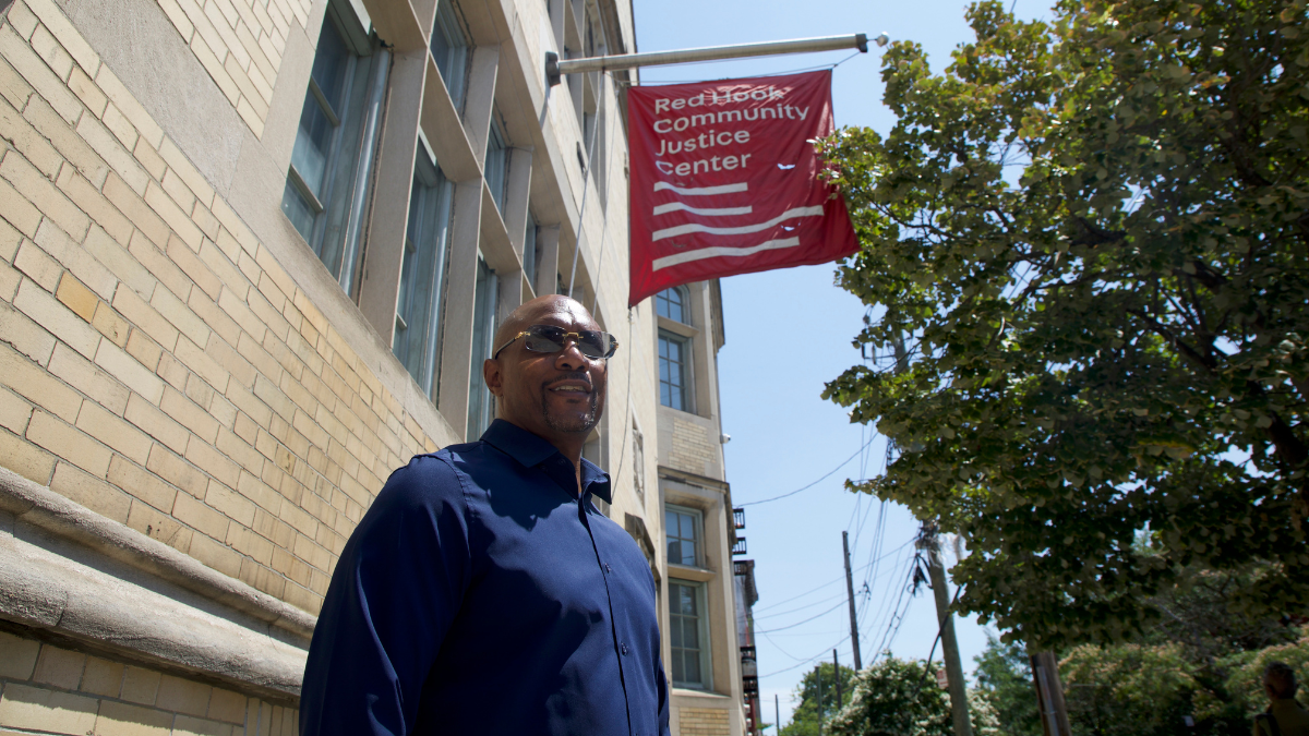 Thaddeus Canty stands with sunglasses and blue button-down shirt underneath a red banner reading "Red Hook Community Justice Center" next to brick building and trees.