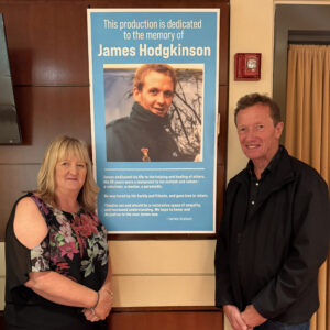 Joan Scourfield and David Hodgkinson stand next to a poster of their son James; bold text on top reads: "This production is dedicated to the memory of James Hodgkinson."