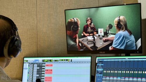 A man sits in a recording studio facing three monitors; on one, two men and one woman are recording a podcast with professional microphones.
