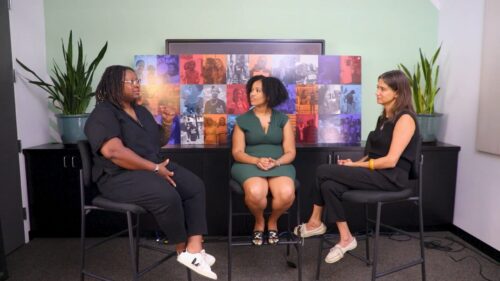 Three women sit in conversation in front of colorful, multi-image display board between two large potted plants.