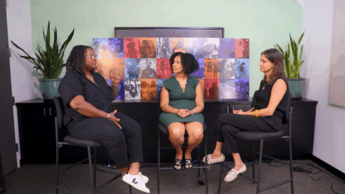 Three women sit in conversation in front of colorful, multi-image display board between two large potted plants.