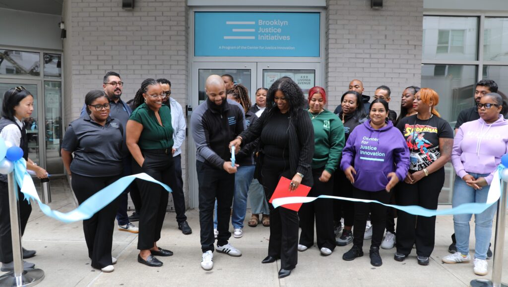 A group of people stand outside brick building with "Brooklyn Justice Initiatives" sign for a ribbon-cutting ceremony at our new Supervised Release office in East New York.