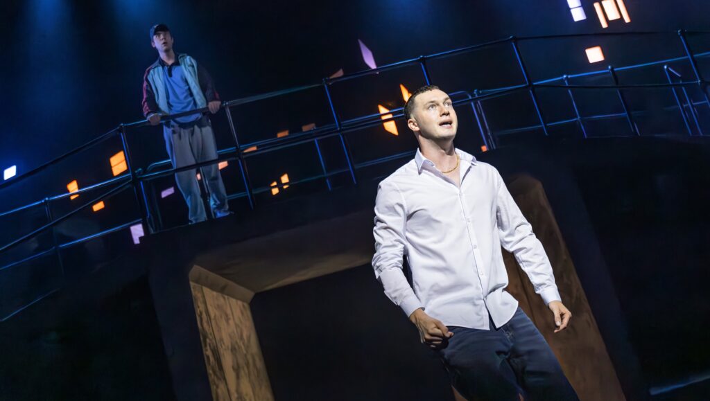 Will Harrison, starring as Jacob Dunne, stands distressed in front of a bridge on stage; Cody Kostro as Raf stands behind and above him.