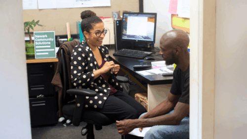 A woman sits smiling at desk next to man leaning forward in his chair; behind her, a green poster reads 
