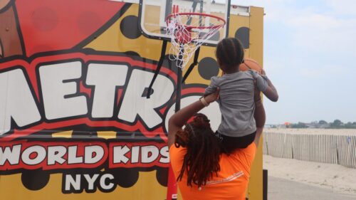 A woman in a Queens Community Justice Center T-shirt lifts a child holding a basketball up on her shoulders towards a hoop.