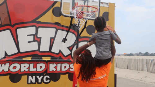 A woman in a Queens Community Justice Center T-shirt lifts a child holding a basketball up on her shoulders towards a hoop.