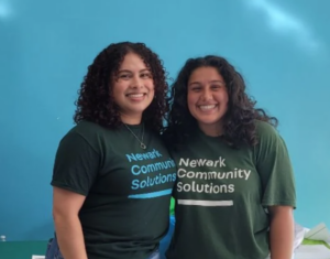 Two women in Newark Community Solutions T-shirts stand smiling in front of blue wall.