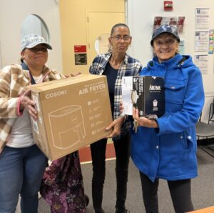 Three women hold up a rice cooker and air fryer in original packaging at our Red Hook Community Justice Center.