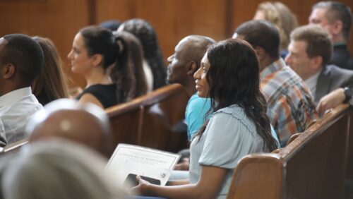 A woman sits smiling at the front of a row of people in a busy courtroom, holding a piece of paper that reads 