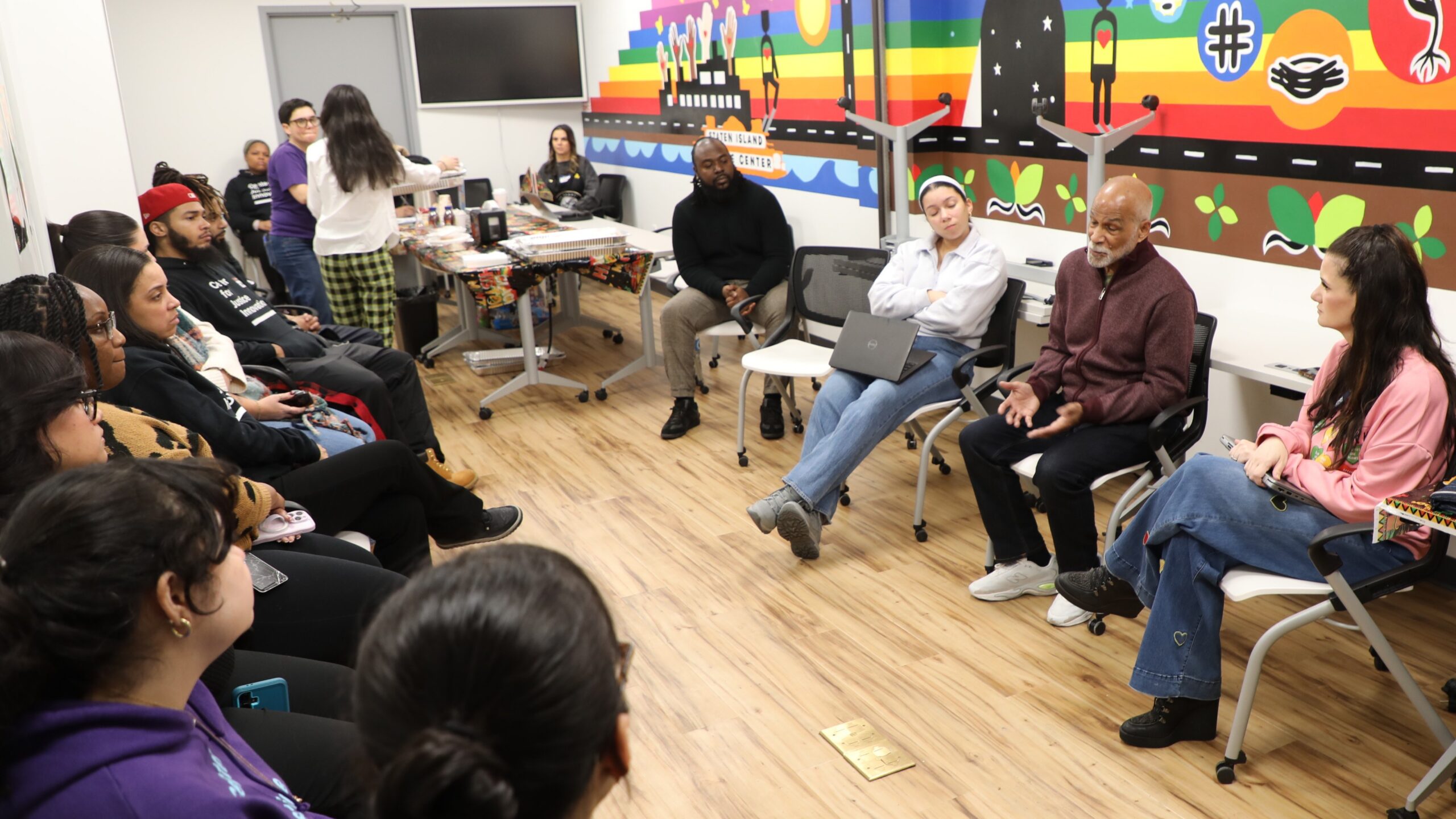 A group of people sit together in the Staten Island Justice Center while one man addresses the group.