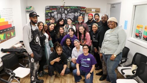 A group of people stand smiling in our Staten Island Justice Center in front of a poster featuring famous Black leaders and historical figures.