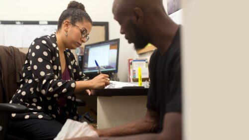A man sits beside a desk, where a woman leans over filling out paperwork next to a desktop monitor.