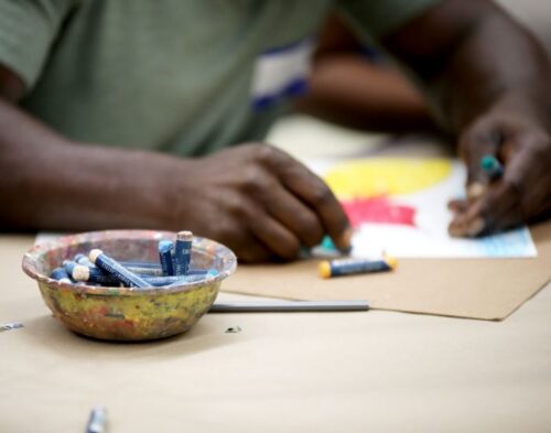 A bowl of oil pastels sits on a table; in the background, the hands of a man drawing on a piece of paper