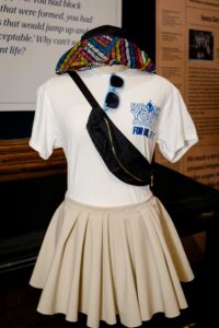 Fashion exhibit featuring sunglasses, T-shirt, crossbody bag, and skirt on a mannequin.