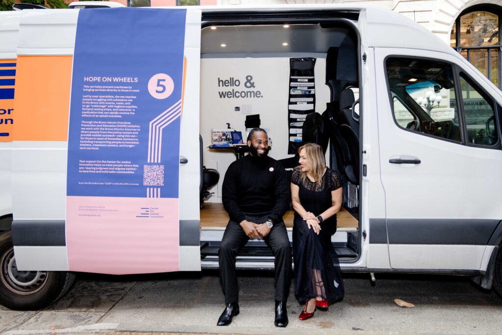A man and woman sit laughing with each other at the entrance of a large white van with a poster on its door that reads "Hope on wheels"; inside, bold text reads "hello and welcome"